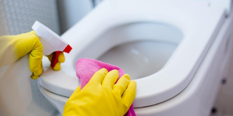 Manos con guantes amarillos limpiando un inodoro blanco con un trapo rosa y un spray.