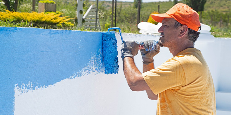 Hombre de perfil pintando la pared de una pileta con pintura celeste.