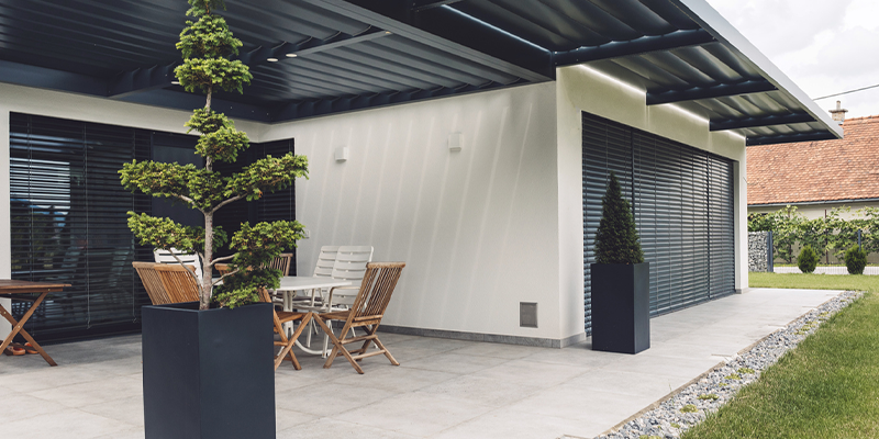 Patio con piso de cerámico blanco, césped, plantas en macetas y juego de sillas y mesa de madera.