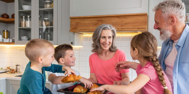 Dos adultos mayores y tres niños comiendo medialunas en la cocina.