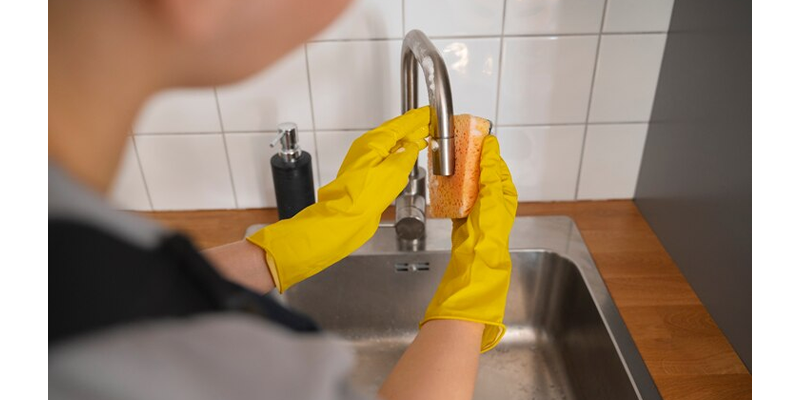 Persona limpiando una canilla, usando guantes amarillos y esponja.